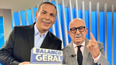 Reinaldo Gottino segura placa do Balanço Geral ao lado de Renato Lombardi com dedo indicador levantado em estúdio da Record