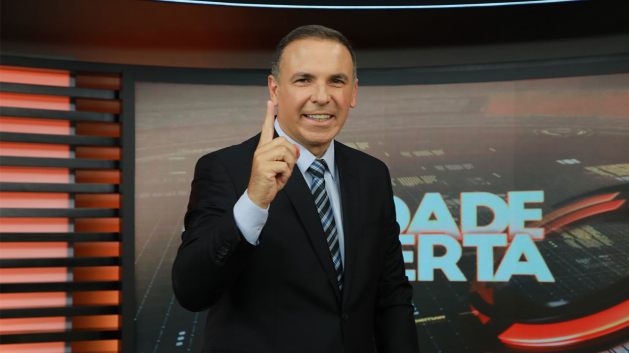 Reinaldo Gottino sorri e aponta o dedo indicador para cima no cenário do Cidade Alerta com logotipo do programa ao fundo na Record