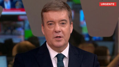 Roberto Kovalick de terno escuro e gravata verde apresenta o telejornal na GloboNews com o selo de urgente exibido no canto da tela