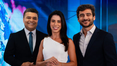 Roberto Nonato, Beatriz Frehner e Evando Cini sorriem em frente ao telão azul do cenário do Jornal da Manhã na Jovem Pan