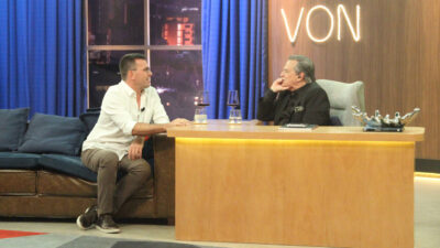 Rodrigo Bocardi de camisa branca conversa com Ronnie Von no cenário do programa Companhia Certa com taças de vinho sobre a mesa na RedeTV!