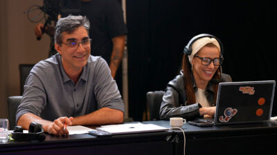 Rodrigo Dourado e Angélica Campos sorriem em mesa de trabalho com notebook do Big Brother Brasil nos bastidores da seleção do reality show