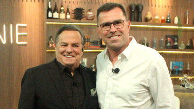 Ronnie Von e Rodrigo Bocardi sorrindo lado a lado no cenário do programa Companhia Certa com prateleiras e objetos decorativos ao fundo