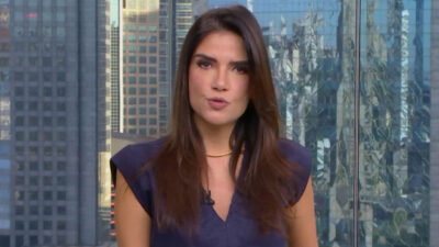 Sabina Simonato de blusa roxa e colar dourado apresenta o Bom Dia São Paulo no estúdio da TV Globo com vista para a cidade ao fundo