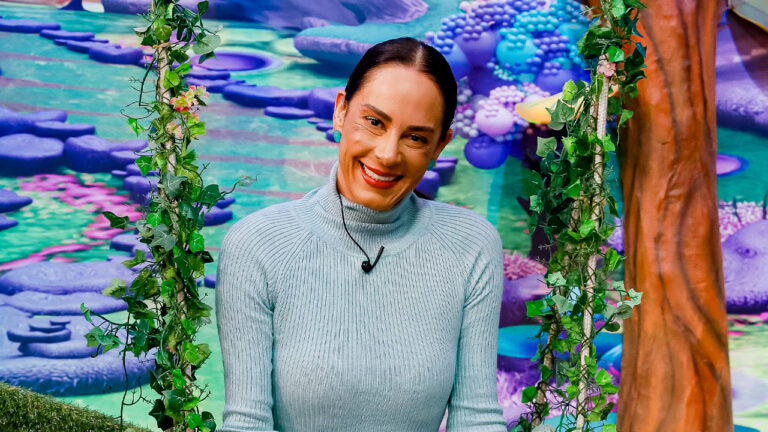 Silvia Abravanel sorri vestindo blusa azul clara em balanço com folhagens no cenário colorido do infantil Sábado Animado no SBT