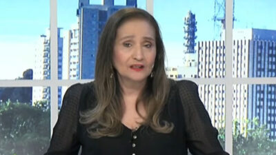 Sonia Abrão com blusa preta de mangas levemente transparentes apresenta o programa A Tarde é Sua diante de janela com vista de prédios