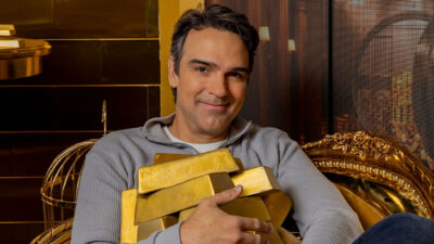 Tadeu Schmidt sorri sentado em poltrona dourada enquanto segura barras de ouro no confessionário do BBB 26 nos estúdios da Globo
