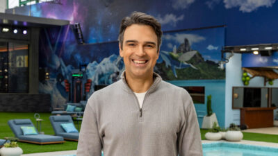 Tadeu Schmidt sorridente com suéter cinza na área externa da casa do BBB 26 com piscina e decoração temática de galáxia ao fundo