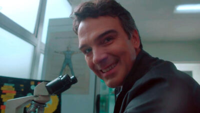 Tadeu Schmidt sorridente ao lado de um microscópio em laboratório durante chamada oficial do BBB 26 na TV Globo