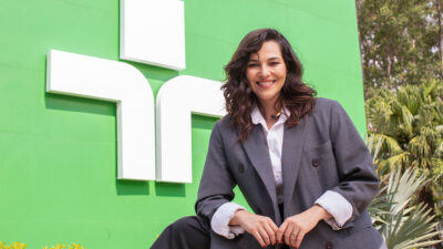 Tainá Müller sorri de blazer cinza e camisa branca à frente do logotipo da TV Cultura em painel verde com vegetação ao fundo em ambiente externo
