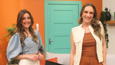 Taisa Pelosi e Paola Pretto sorriem lado a lado no estúdio do Manhã Com Você da RedeTV! com parede laranja e porta verde atrás