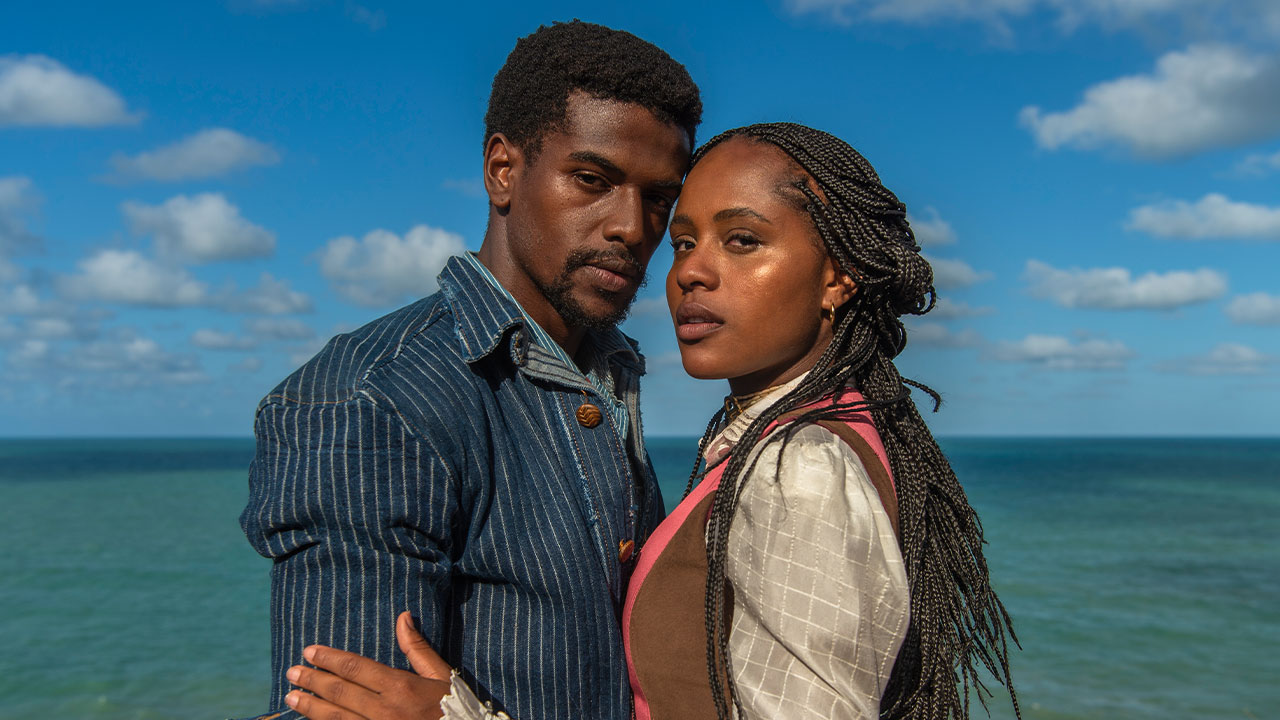 Tonho (Ronald Sotto) e Alika (Duda Santos) abraçados à beira do mar em cena de época da novela A Nobreza do Amor