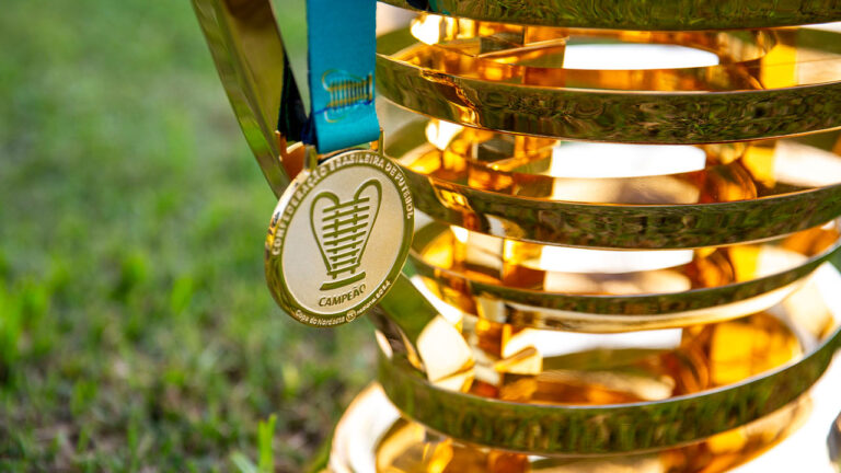 Medalha dourada de campeão pendurada na taça da Copa do Nordeste formada por anéis dourados e gramado verde desfocado ao fundo
