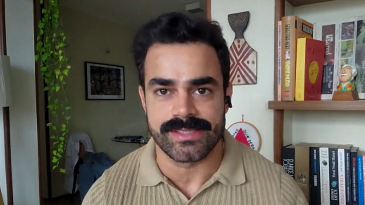 Túlio Amâncio fala em vídeo com fone no ouvido em ambiente doméstico com estante de livros e objetos decorativos ao fundo