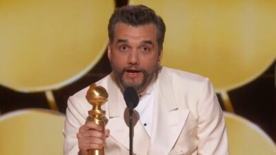 Wagner Moura segura o troféu do Globo de Ouro usando terno branco enquanto discursa no palco da premiação internacional de cinema