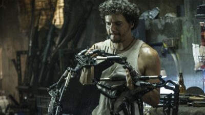 Wagner Moura como Spider segura um exoesqueleto mecânico em oficina no filme Elysium com expressão concentrada e armas ao fundo