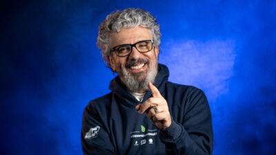 Zé Luiz sorri apontando o dedo indicador em retrato de estúdio com moletom escuro óculos e fundo azul iluminado