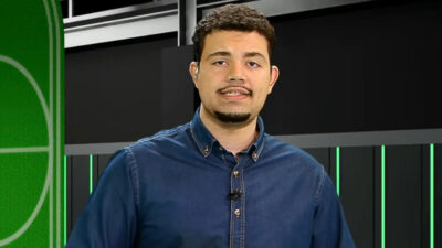 Zé Neto narrador da Xsports com camisa jeans azul e microfone de lapela fala durante transmissão esportiva em estúdio virtual