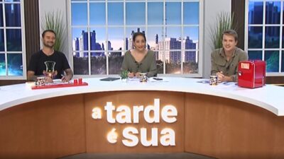 Alessandro Lo-Bianco, Sonia Abrão e Aaron Tura sorriem na bancada do programa A Tarde é Sua, estúdio com logo da atração e vista de cidade ao fundo