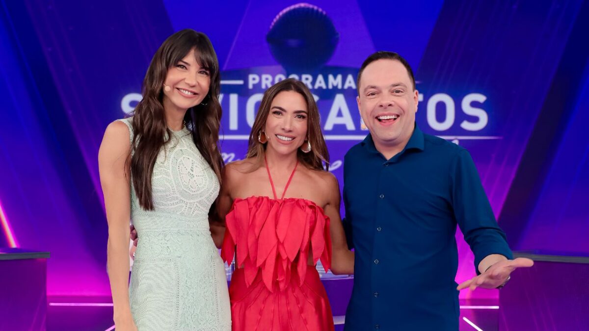 Amanda Klein, Patricia Abravanel e Thiago Gardinali sorriem no palco do Programa Silvio Santos, vestindo roupas clara, vermelha e azul escura, no SBT