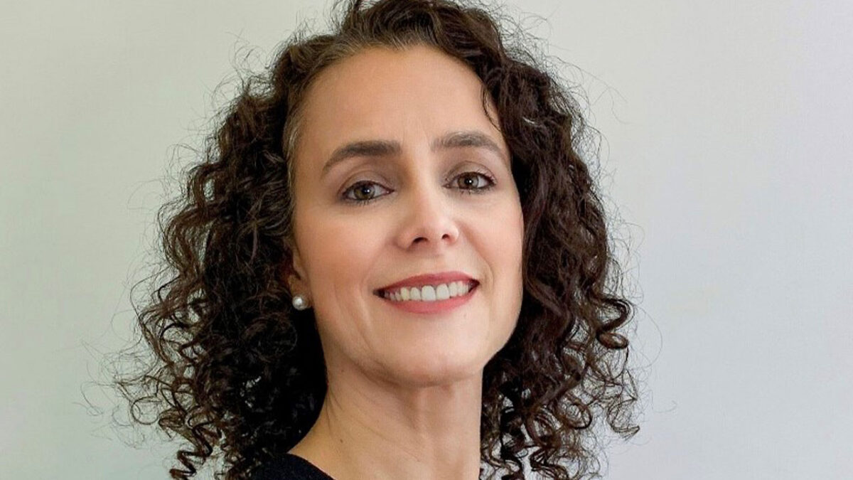 Ana Eliza Faria e Silva sorri para a foto exibindo cabelos castanhos cacheados e volumosos com brincos de pérola discretos sobre um fundo cinza claro