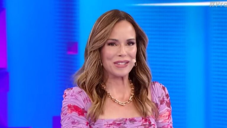 Ana Furtado sorri em estúdio de TV, veste blusa floral lilás e rosa com decote quadrado, usa colar de corrente dourada e tem cabelos castanhos soltos