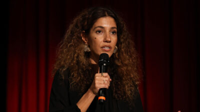 Antonia Pellegrino, diretora da EBC, fala ao microfone em evento, mulher de cabelos cacheados volumosos, veste blusa preta, usa brincos e fundo vermelho