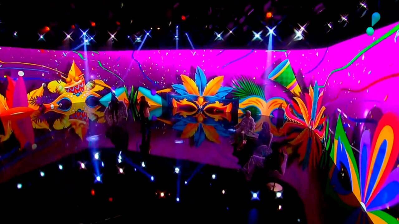 Cenário do Bastidores do Carnaval da RedeTV! com painéis de LED em tons de roxo e rosa, exibe máscaras de Carnaval coloridas e luzes de estúdio, refletidas no chão preto brilhante