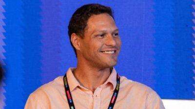 Ben-Hur Correia sorri usando camisa social clara e crachá no pescoço diante de painel azul em evento
