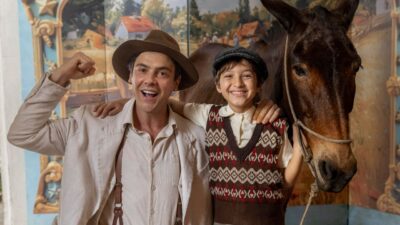 Candinho (Sergio Guizé) e Samir (Davi Malizia) sorriem abraçados ao lado de um burro, vestem figurinos de época da novela Êta Mundo Melhor!, na TV Globo