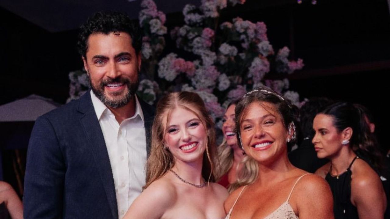 Carlo Porto, Lorena Queiroz e Pri Sol posam sorrindo juntos em evento com decoração floral ao fundo e convidados ao redor