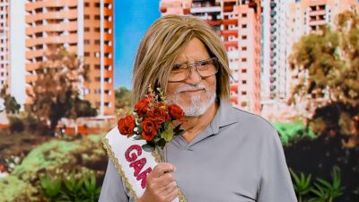 Carlos Alberto de Nóbrega usa peruca loira, óculos e barba branca, segura buquê de rosas vermelhas e veste faixa de miss em cenário de prédios coloridos