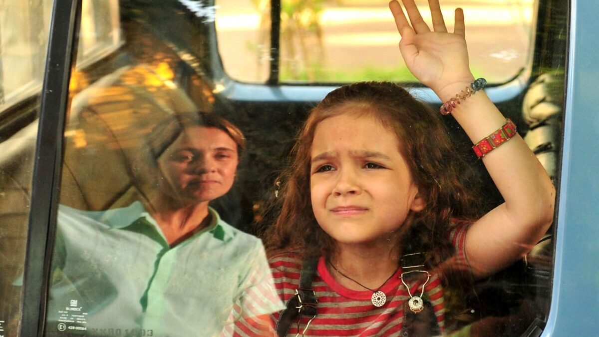 Carminha interpretada por Adriana Esteves observa Rita vivida por Mel Maia pela janela do carro enquanto a menina levanta a mão com expressão tensa em cena de Avenida Brasil
