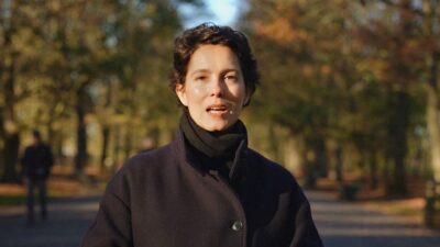 Cecília Malan de cabelos curtos castanhos usa casaco azul marinho e gola alta preta enquanto fala em ambiente externo com árvores ao fundo no outono