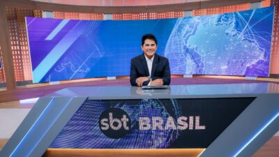 César Filho sorri sentado na bancada do telejornal SBT Brasil, veste terno escuro e camisa branca, ao fundo telão azul com mapa mundi tecnológico