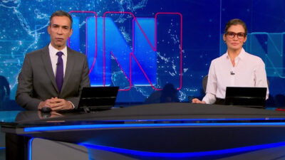 Apresentadores César Tralli e Renata Vasconcellos na bancada do Jornal Nacional com cenário azul ao fundo exibindo mapa-múndi e logotipo do telejornal