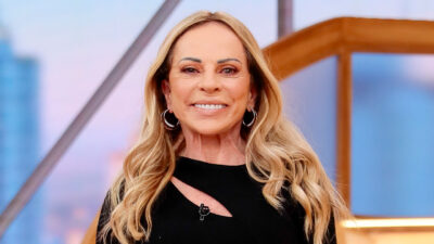 A apresentadora Christina Rocha sorri para a câmera usando um vestido preto com decote assimétrico e brincos de argola em um cenário iluminado