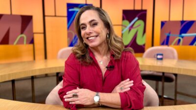 Cissa Guimarães sorri de braços cruzados em uma bancada de madeira, vestindo camisa vermelha e relógio prateado, no cenário do programa Sem Censura