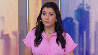 Apresentadora Dani Brandi de blusa rosa e colar prateado olha para a câmera nos estúdios do programa Alô Você com cenário digital azul ao fundo