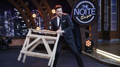 Danilo Gentili veste terno escuro e inclina o corpo para frente enquanto segura um cavalete de madeira no palco do programa The Noite, no SBT