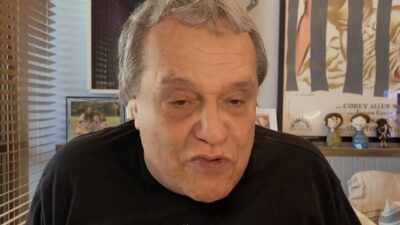 Dennis Carvalho fala de frente, usando fones de ouvido sem fio e camiseta preta, em ambiente doméstico com persiana, quadros e bonecos ao fundo