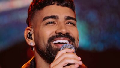 Cantor Dilsinho sorri enquanto segura microfone próximo ao rosto durante apresentação, usando fone de retorno, brinco brilhante e barba aparada