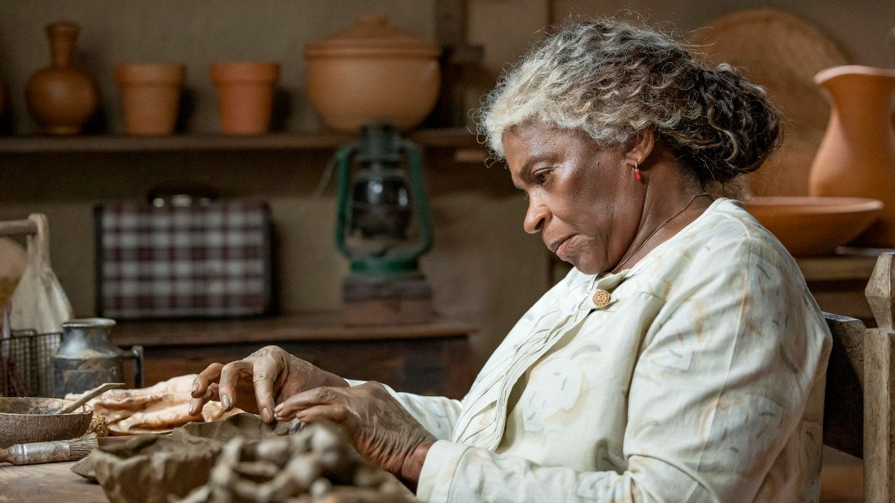 Zezé Motta como Dona Menina em A Nobreza do Amor, moldando argila com as mãos em oficina rústica com vasos de barro e lampião ao fundo da cena
