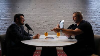 Dr Adriano Almeida e Rodrigo Alvarez sentados frente a frente no programa Rodrigo Alvarez Entrevista com microfones sobre mesa redonda e parede de tijolos ao fundo