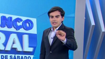 Apresentador Dudu Camargo veste terno preto e camisa clara, gesticula com a mão esquerda à frente de telão azul com logotipo do programa Balanço Geral