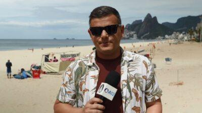 Repórter Eduardo Gois, de óculos escuros e camisa florida, segura microfone da TV Aparecida no Arquivo A, com a praia e o Morro Dois Irmãos ao fundo