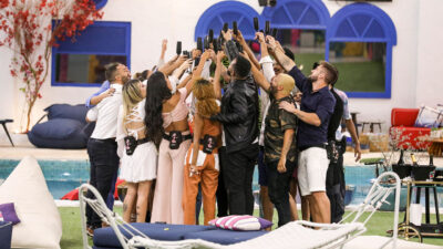 Participantes do BBB 21 reunidos em círculo na área externa da casa, brindando com taças pretas em celebração, ao lado da piscina e jardim da casa