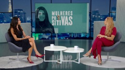 Fabi Saad e Hortência Marcari sentadas em poltronas cinzas num estúdio de TV com painel ao fundo escrito Mulheres Positivas e vista para a cidade