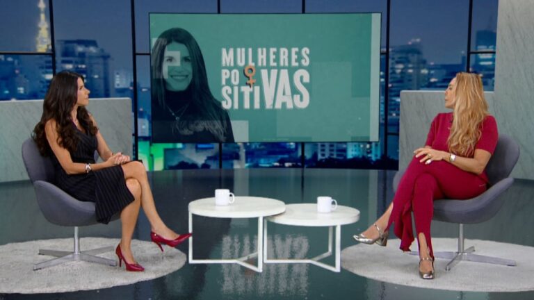 Fabi Saad e Hortência Marcari sentadas em poltronas cinzas num estúdio de TV com painel ao fundo escrito Mulheres Positivas e vista para a cidade