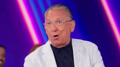 Galvão Bueno sorri com expressão de surpresa, usando óculos de grau e paletó cinza claro sobre camisa polo azul escura, em cenário roxo vibrante